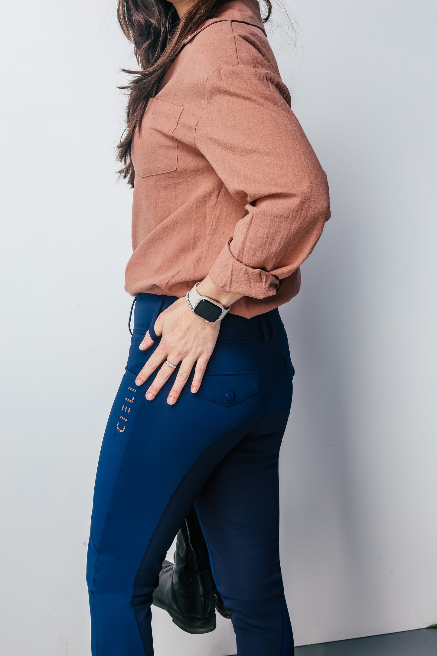 Person wearing a brown shirt and blue riding pants with a visible brand logo, standing against a plain background.