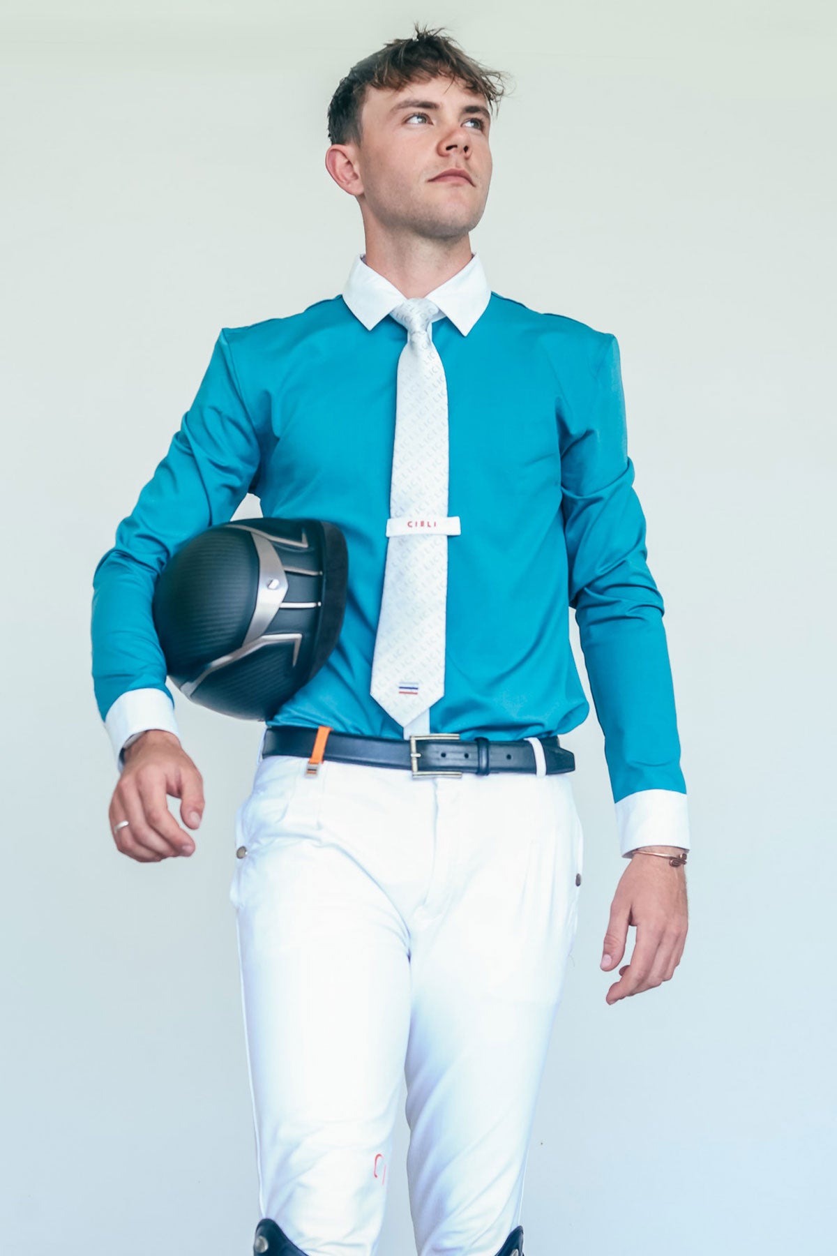 Man in equestrian attire holding a helmet against a plain background