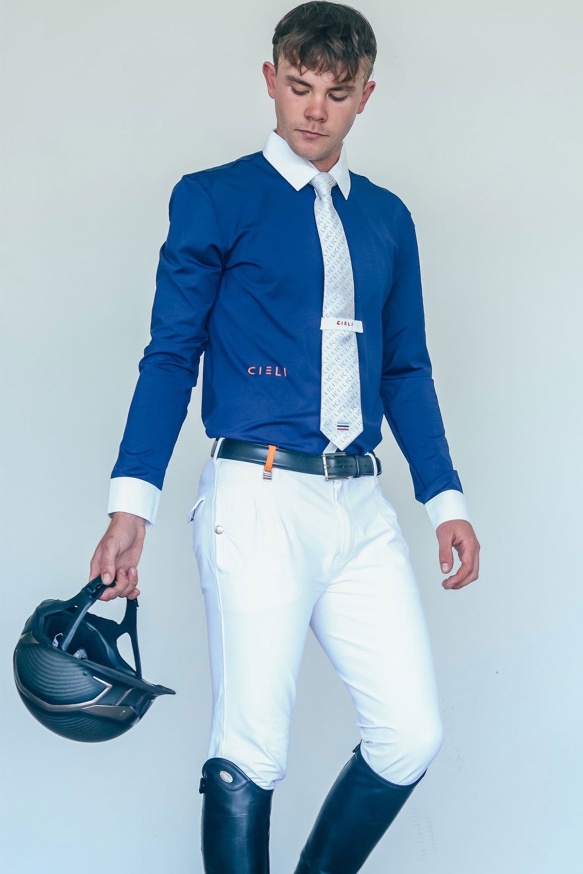Man in equestrian attire holding a helmet against a plain background