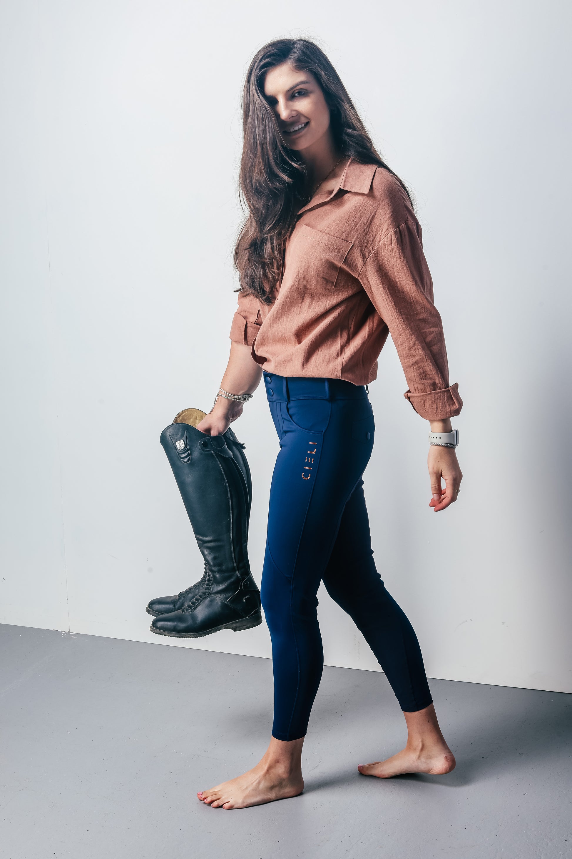 Woman holding riding boots, wearing a brown shirt and dark blue riding pants against a white background