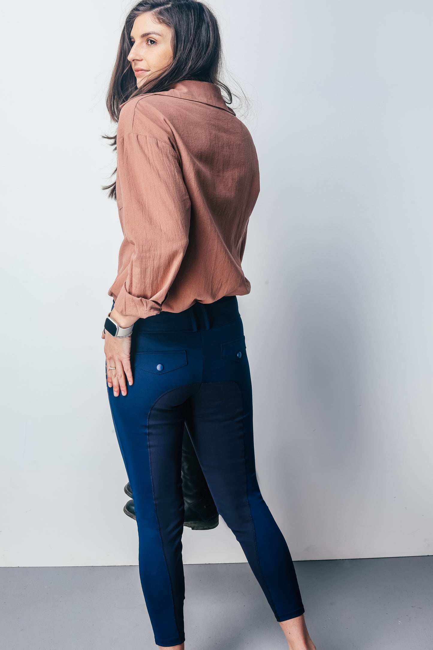 Woman wearing a brown shirt and blue riding pants against a plain background