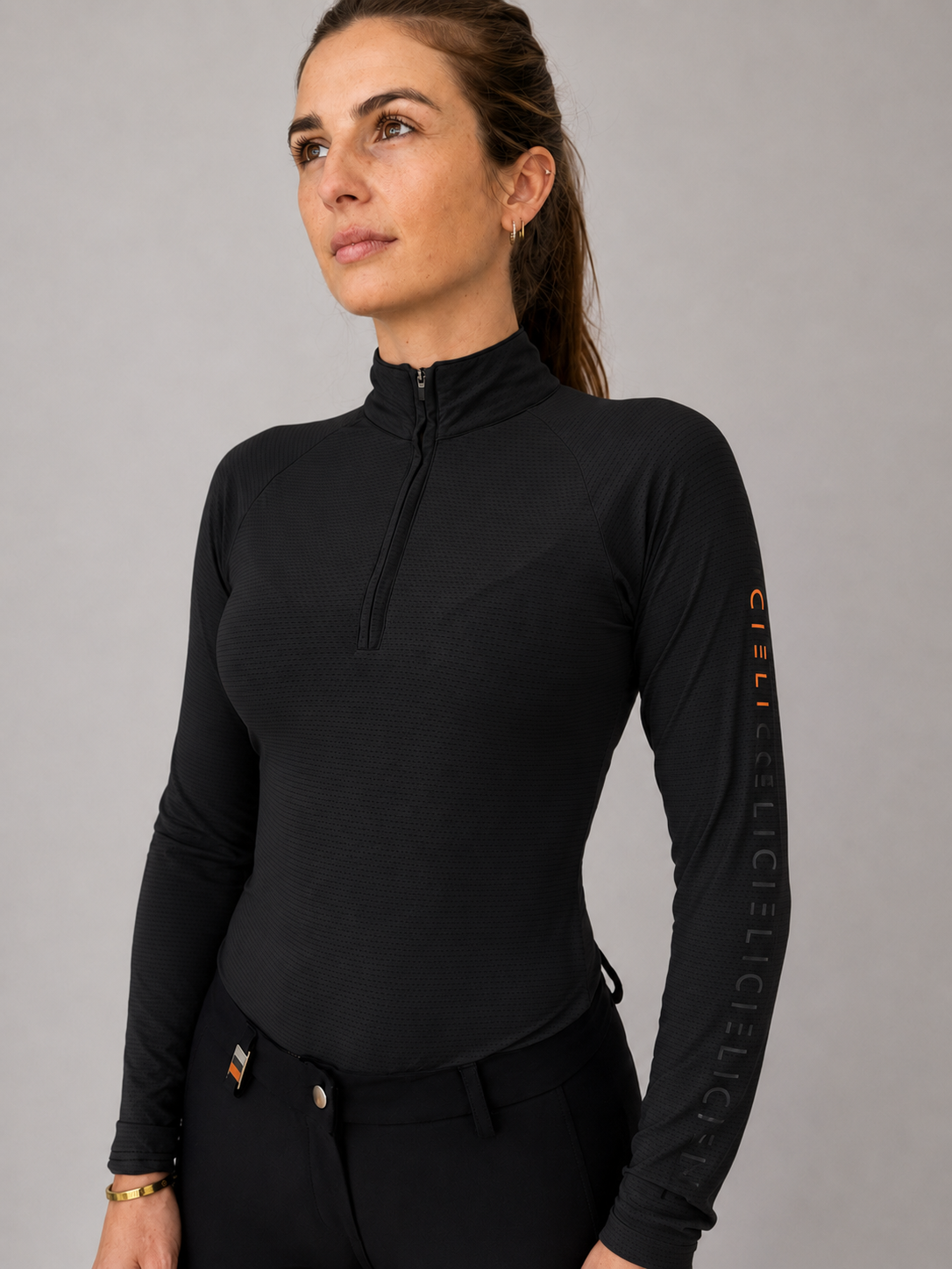 A woman wearing a black long-sleeve half-zip athletic top with vertical 'CIELI' branding on the right sleeve, paired with black pants, against a neutral gray studio background.