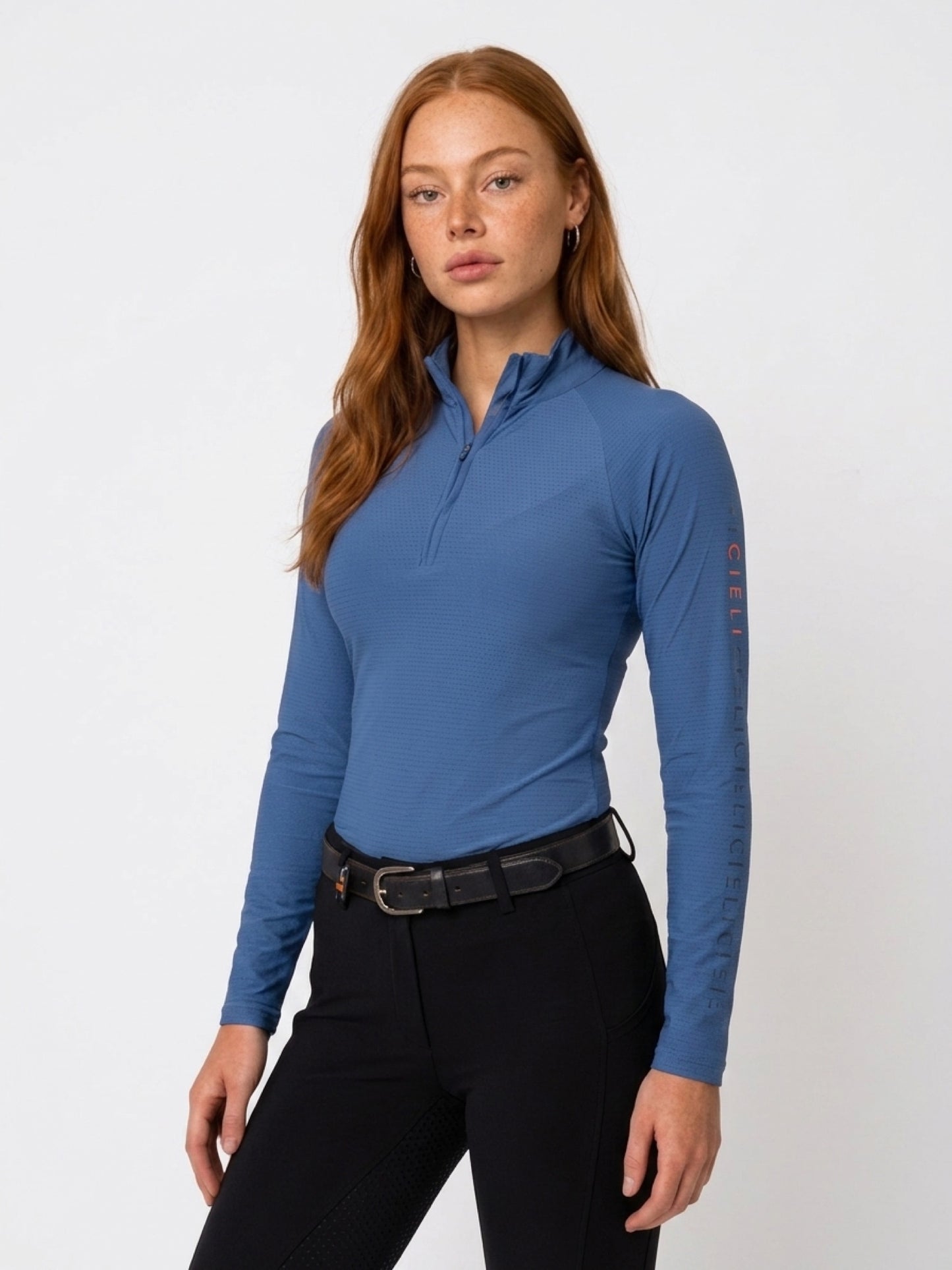 A woman modeling a long-sleeve quarter-zip athletic top in slate blue, paired with black riding pants and a black leather belt, against a plain white studio background.