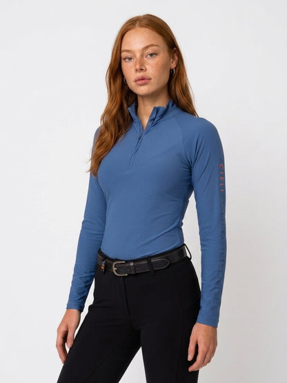 A woman modeling a long-sleeve quarter-zip athletic top in slate blue, paired with black riding pants and a black leather belt, against a plain white studio background.