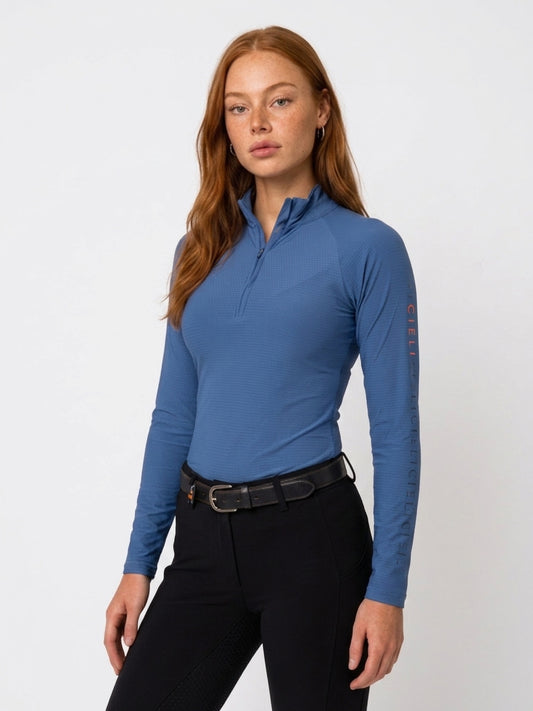 A woman modeling a long-sleeve quarter-zip athletic top in slate blue, paired with black riding pants and a black leather belt, against a plain white studio background.