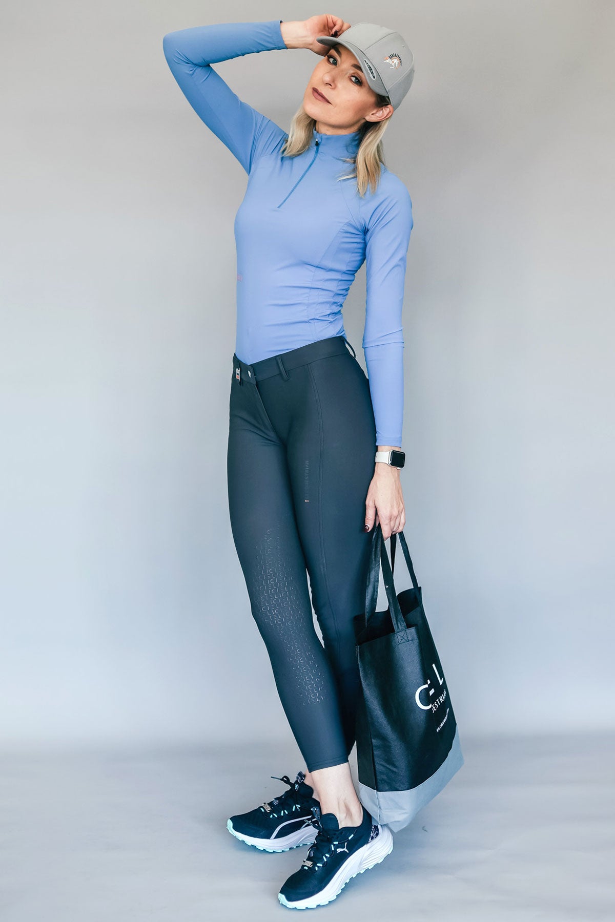 Woman wearing a blue long-sleeve top and black breeches, holding a black bag with a logo.