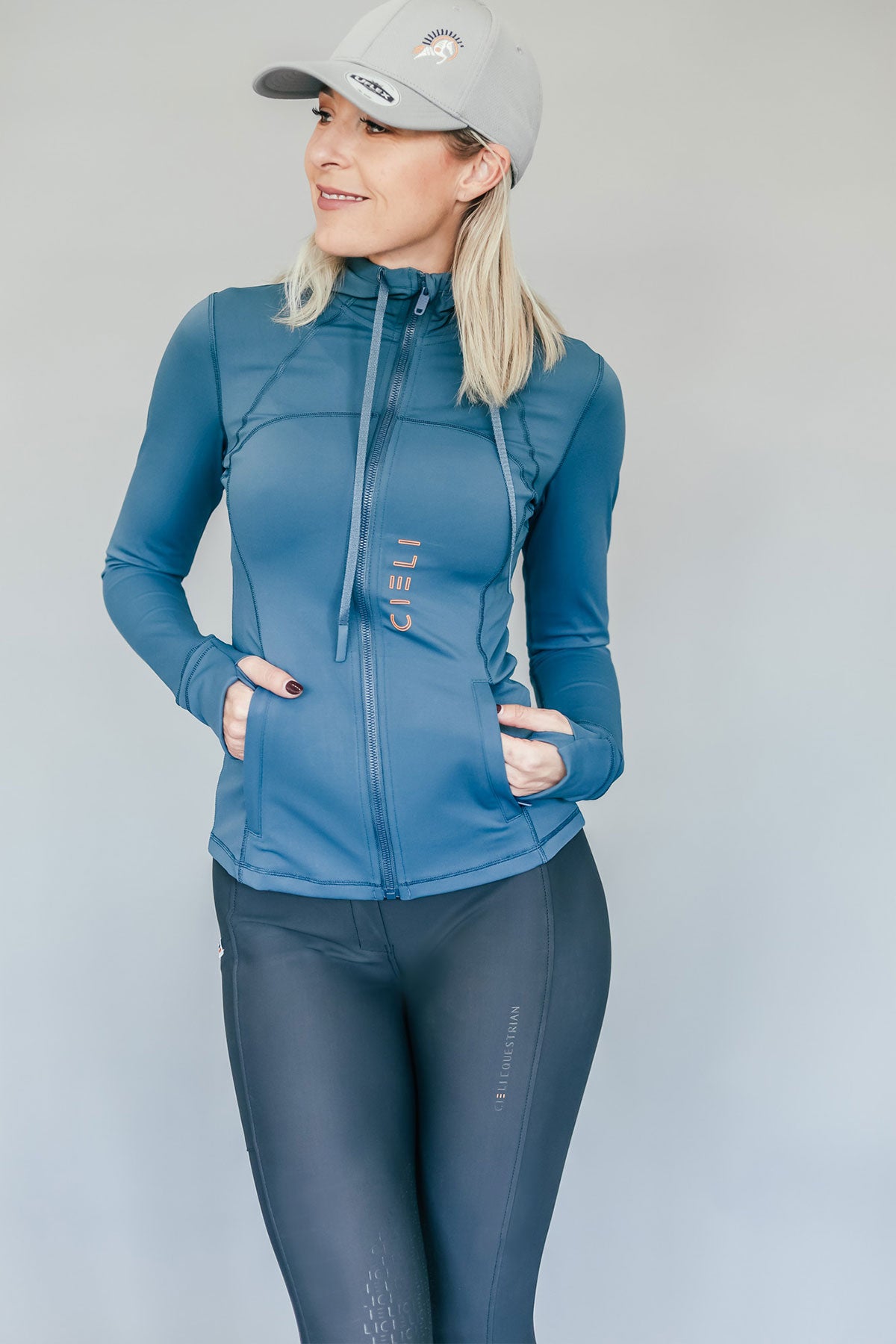 Woman wearing a blue athletic jacket and gray riding pants against a plain background