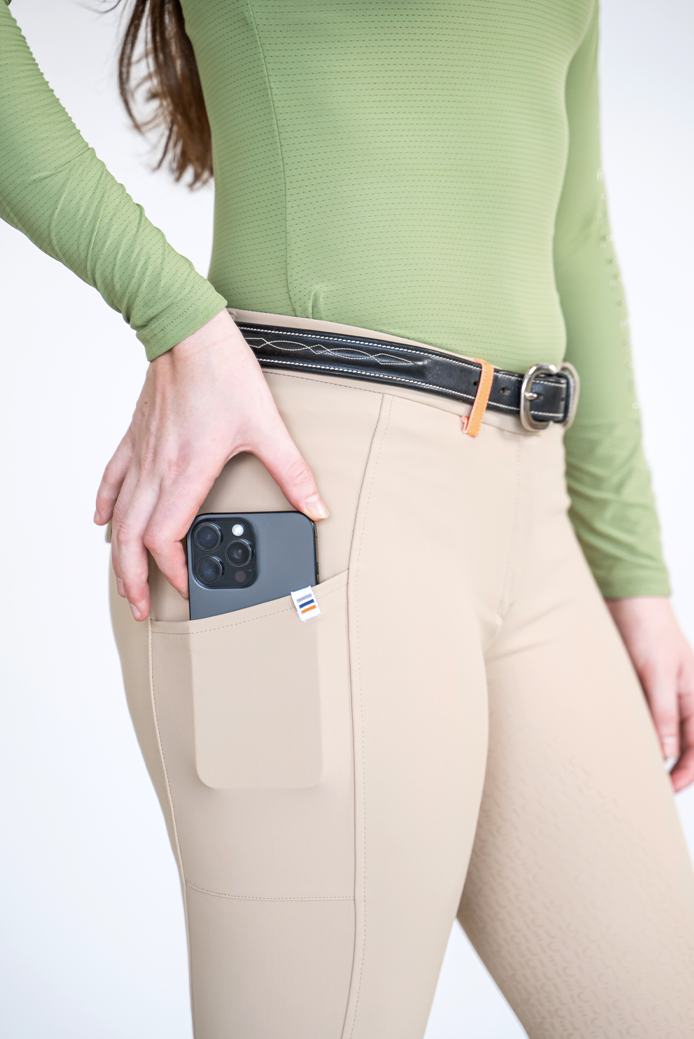 Beige riding breeches with a side pocket holding a smartphone, shown on a model wearing a green ribbed long-sleeve top and a black belt.