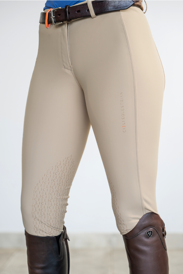 Beige equestrian riding breeches with textured knee patches and a subtle vertical logo, shown on a model wearing a dark brown leather belt and tall dark brown riding boots against a light gray studio background.