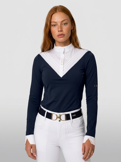 A woman modeling a navy blue and white long-sleeve equestrian shirt with a high mock neck and a white V-neck panel featuring vertical pleats and a small gold logo. She is wearing white riding pants and a black belt with a gold D-ring buckle.