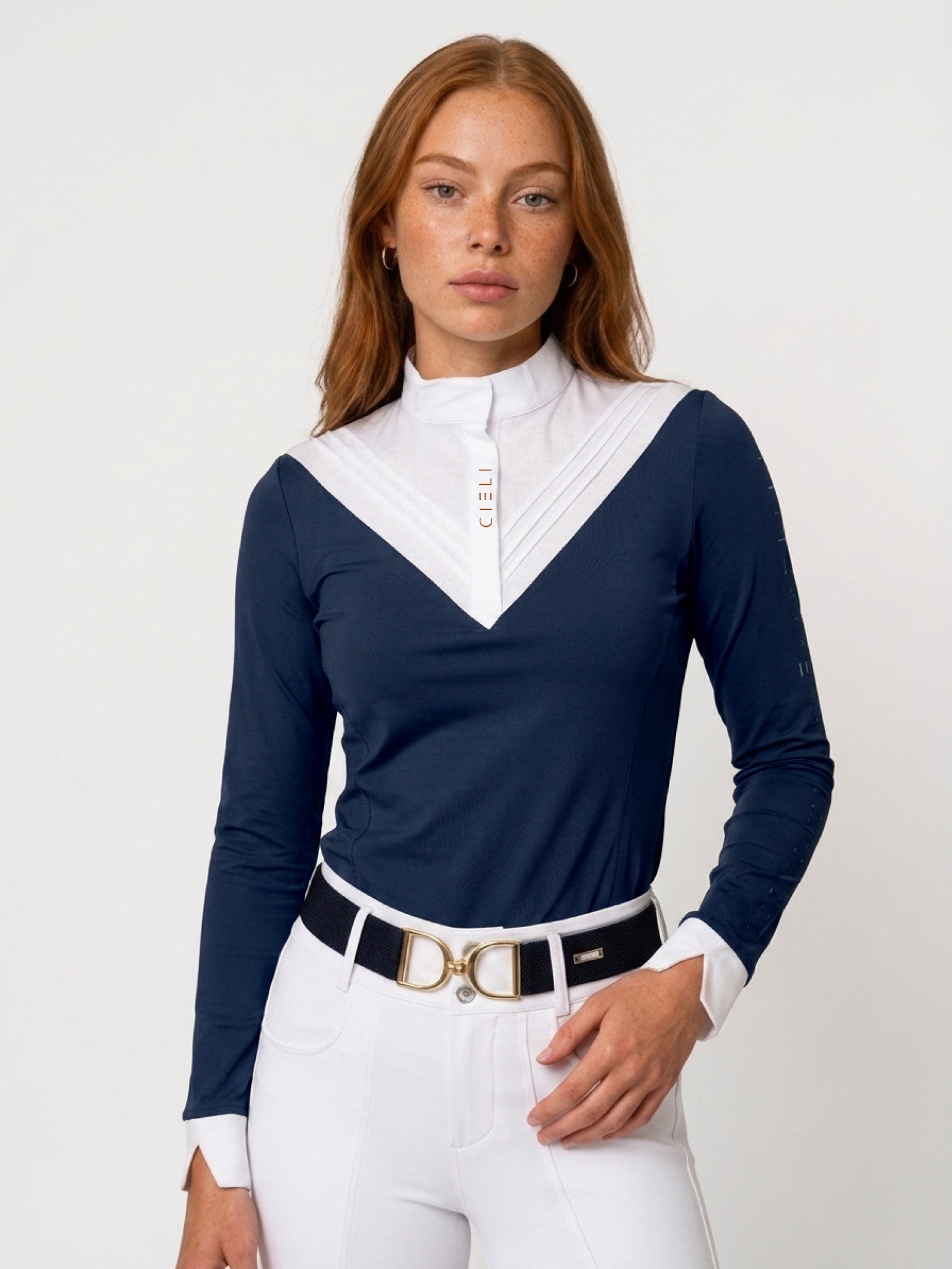 A woman modeling a navy blue long-sleeve polo shirt with a contrasting white pleated V-neck collar, paired with white riding breeches and a black belt with a gold buckle, against a plain white studio background.