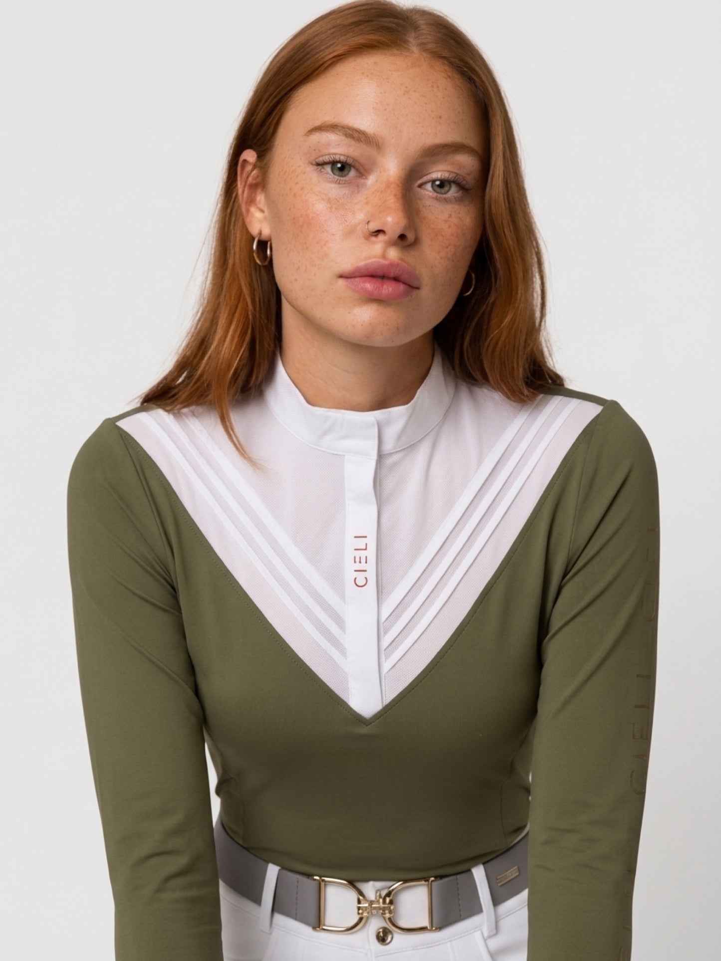 A woman wearing a long-sleeve olive green top with a white pleated V-neck collar and a white front placket featuring the brand name 'CIELI' in red. She is also wearing a white top and a gray belt with a gold buckle, photographed against a light gray studio background.