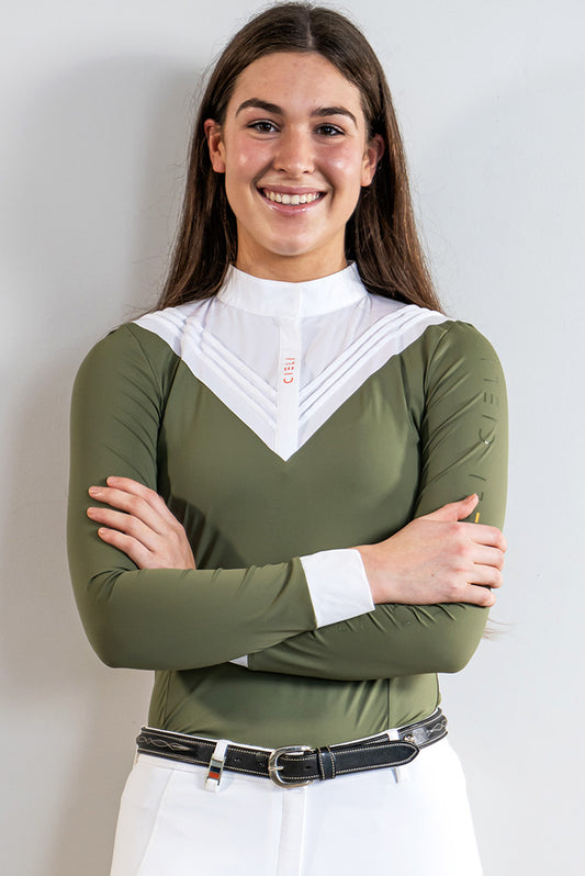 A smiling young woman with long brown hair stands with arms crossed, wearing an olive green long-sleeve riding shirt with a white V-neck collar and white riding pants against a light gray background.