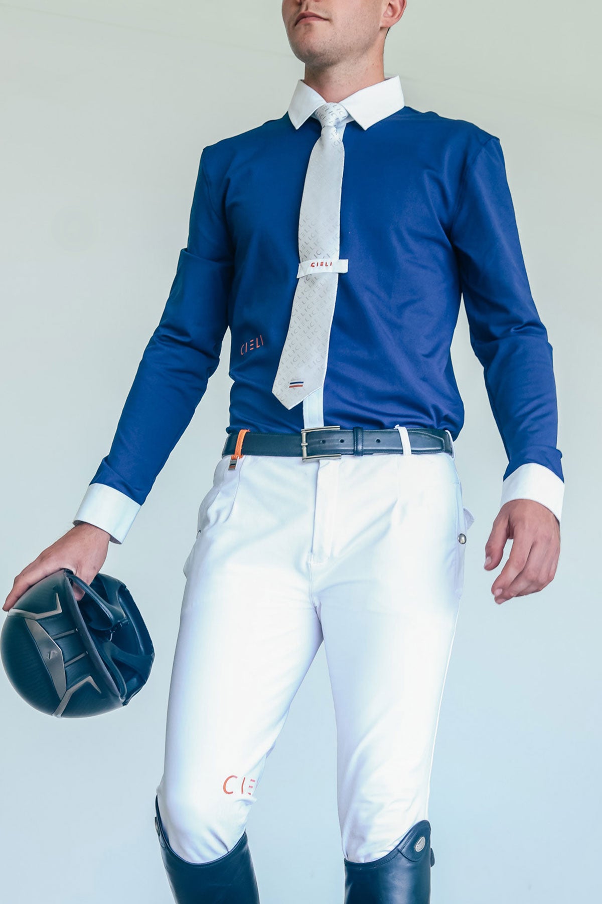 Man wearing a blue shirt, white pants, and riding boots holding a helmet against a light gray background