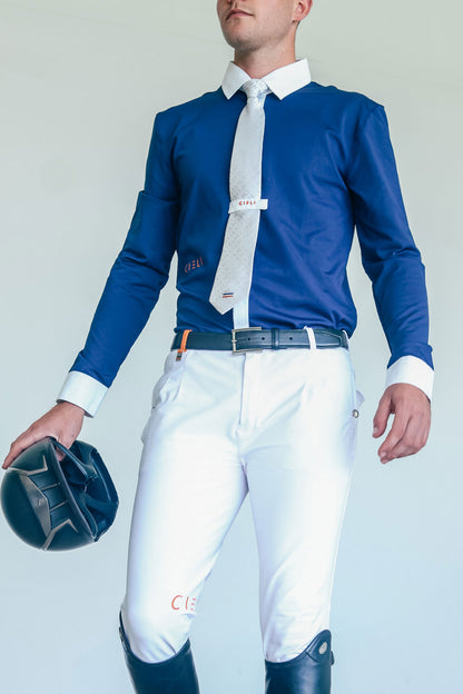 Man wearing a blue shirt, white pants, and riding boots holding a helmet against a light gray background