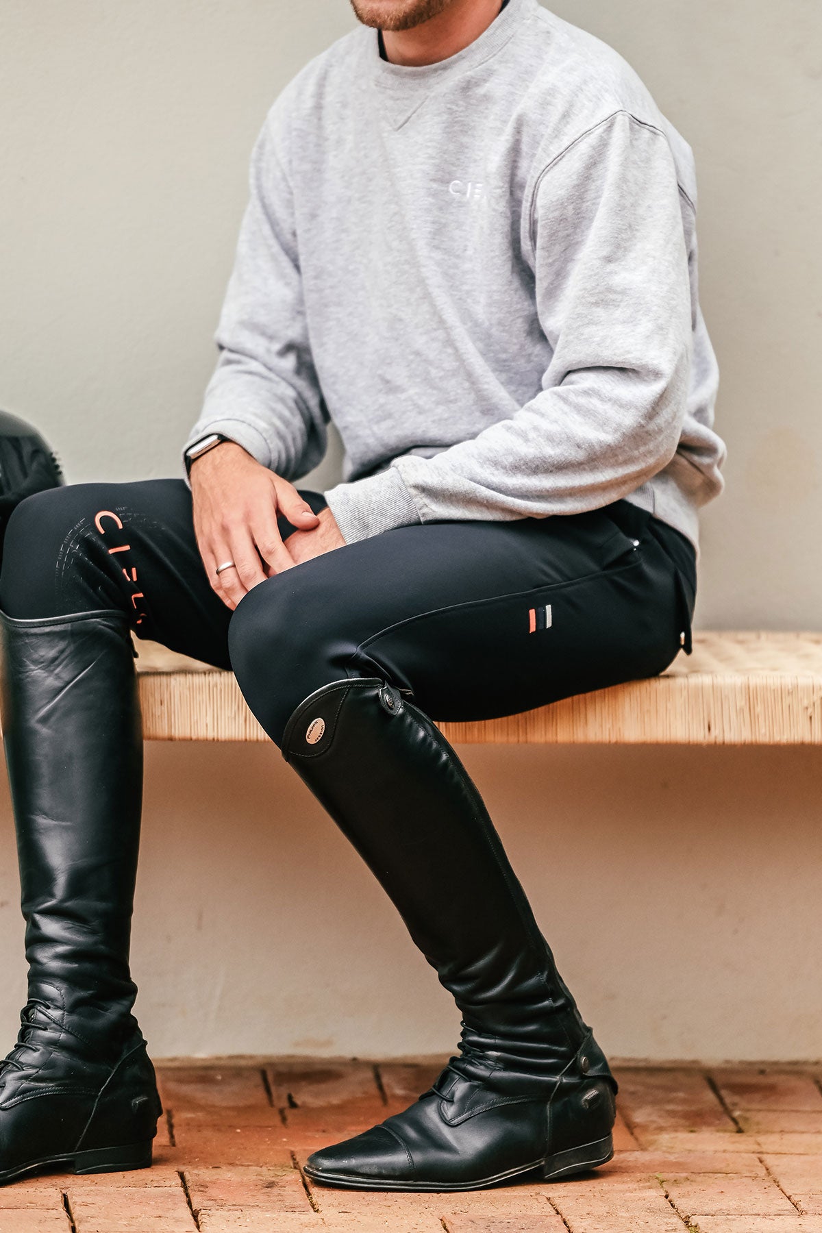 Person wearing black riding boots and pants sitting on a wooden bench.