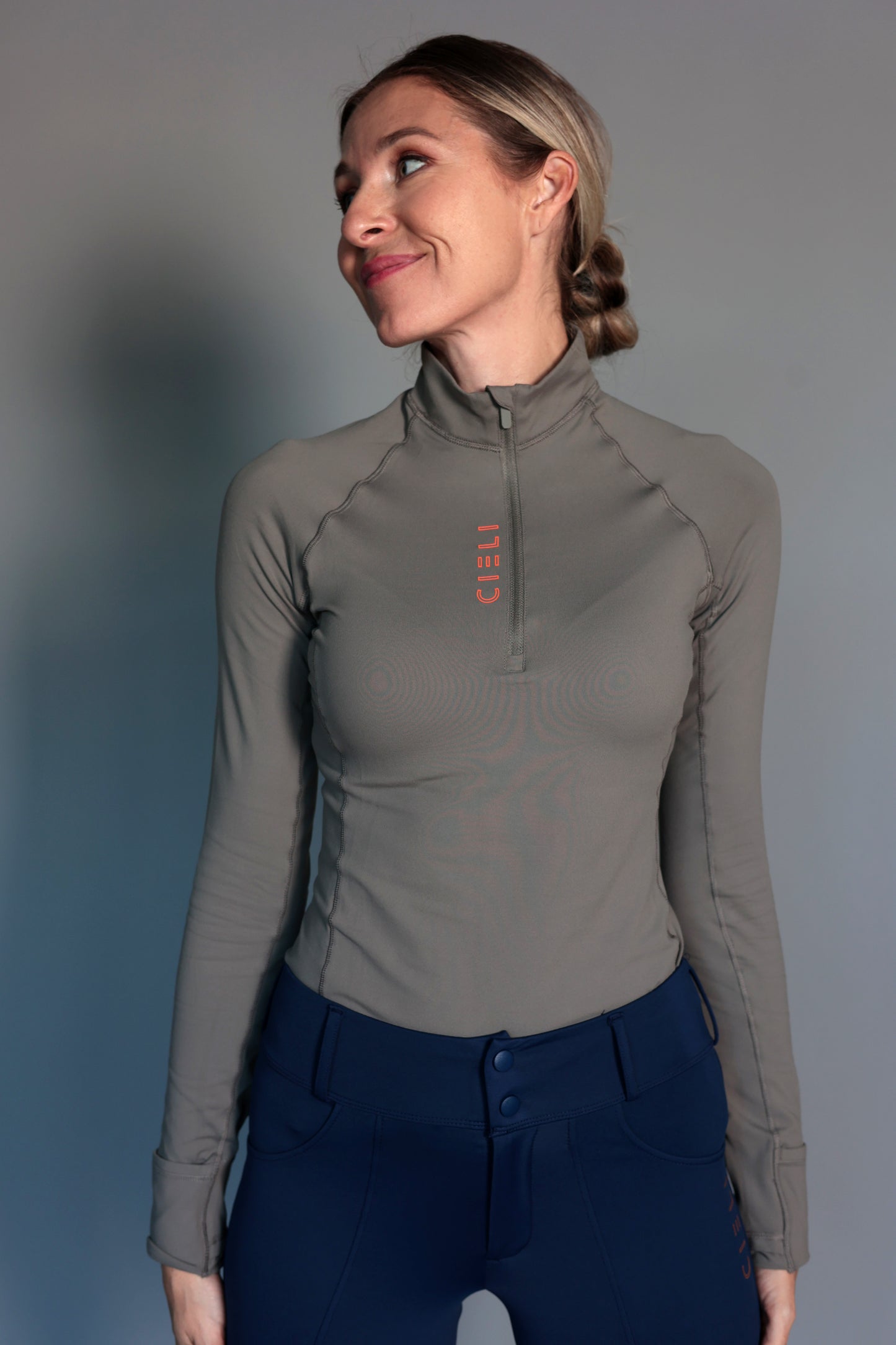 Woman wearing a green equestrian long-sleeve athletic top with a zipper and navy breeches against a gray background