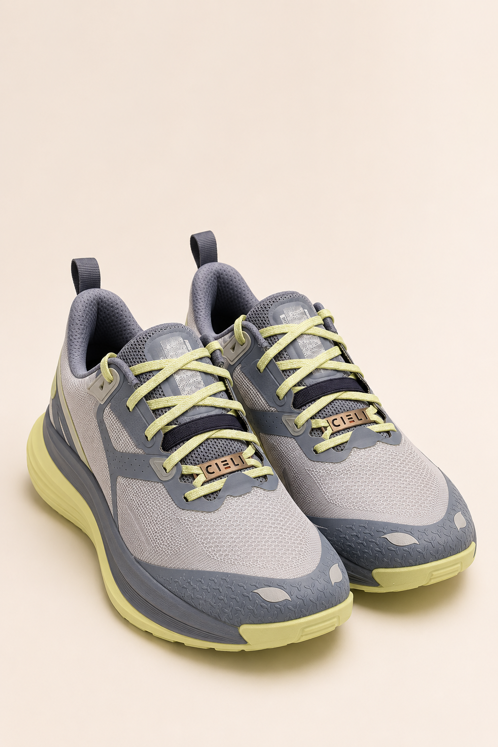 A pair of athletic sneakers in light gray and slate blue-gray with neon yellow-green accents on the laces, logo tab, and sole, displayed on a solid beige background.