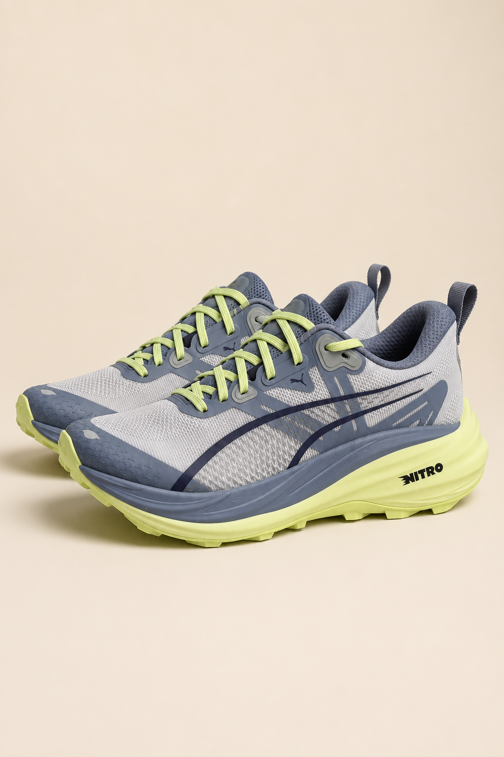 A pair of running shoes in light gray and slate blue with neon yellow-green laces and midsole accents, shown against a solid beige background.