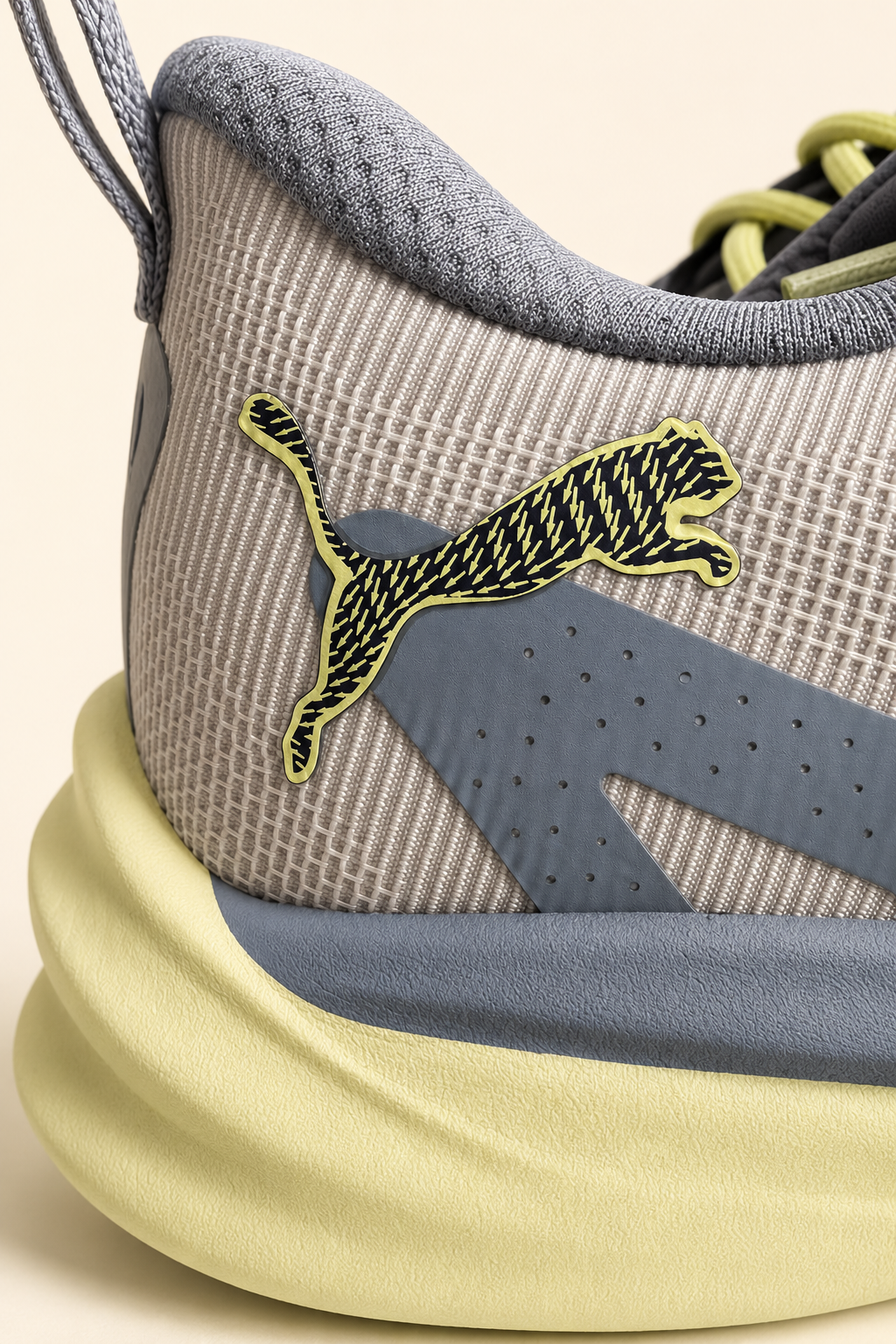Close-up of a Puma sneaker showing the heel area with a light gray knit upper, a gray side stripe, a yellow-green Puma cat logo, and a pale yellow sole on a warm beige background.