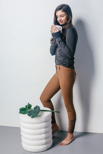 A woman modeling a dark gray long-sleeve athletic top and brown riding breeches, holding a white mug. She is standing next to a white textured stool with green leaves against a light gray background.
